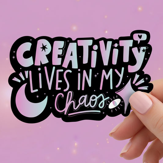 Hand holding a sticker with 'Creativity lives in my chaos' text on a pink background