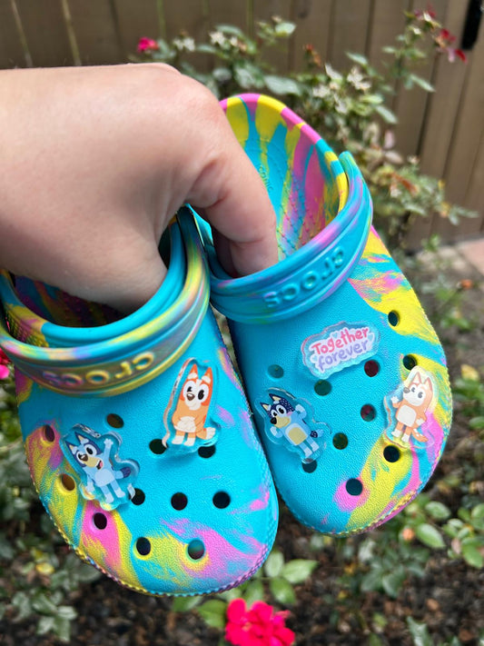 Colorful children's clogs with cartoon characters held by a hand against a garden background.