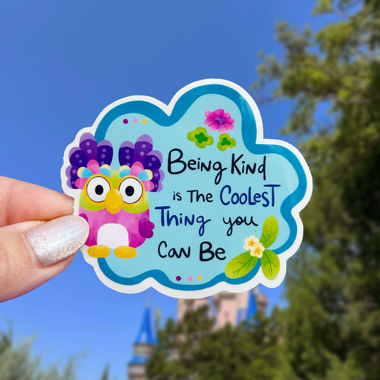 Colorful sticker with owl design and text 'Being Kind is The Coolest Thing you Can Be' held by a hand against a blurred outdoor background.